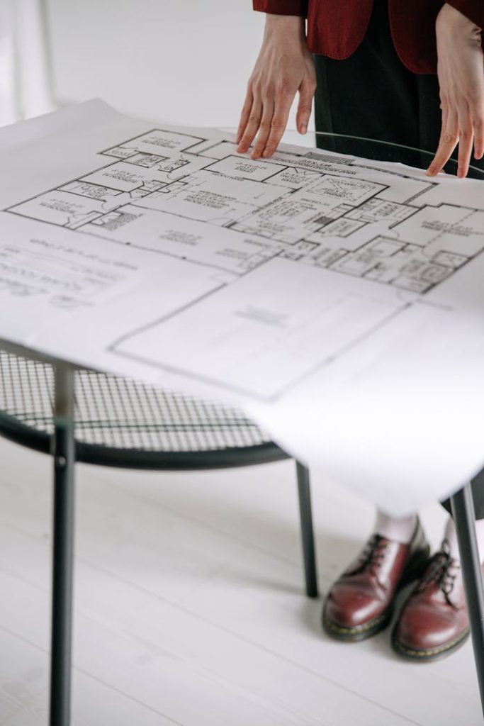 a-person-holding-a-blueprint-8962797 Person examining a detailed architectural floor plan on a table indoors with a focus on design.